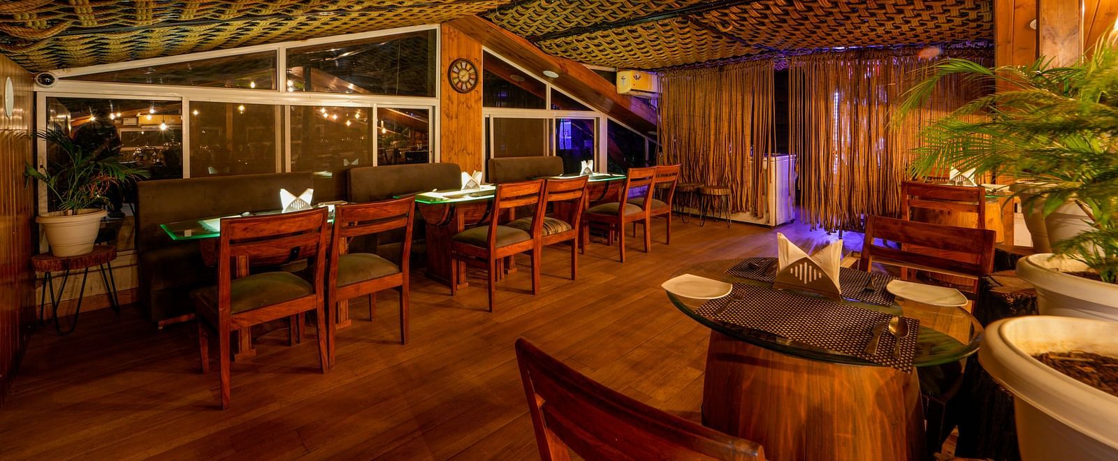 A shot taken from an angle of Ruttba which has wooden interiors and chairs and tables with bright lighting - DLS Whispering Winds