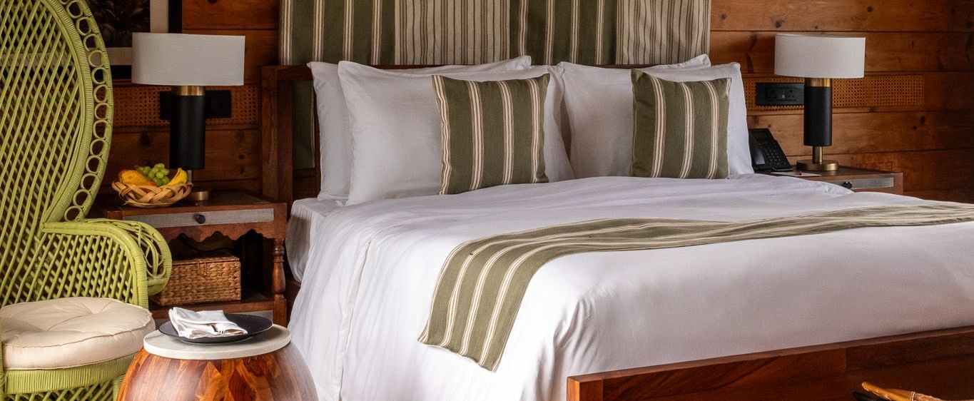 A vertical shot of a bedroom with a king-sized bed, a striped green and white canopy, and a large green wicker chair. A wooden side table and a large framed black and white photo on the wooden paneled wall complete the rustic, yet elegant look. This is a r