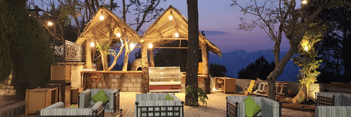 Outdoor seating area at dusk with cushioned chairs and sofas, illuminated by hanging lights under thatched-roof structures, surrounded by trees and a scenic mountain view in the background - Suryavilas Luxury Resort and Spa