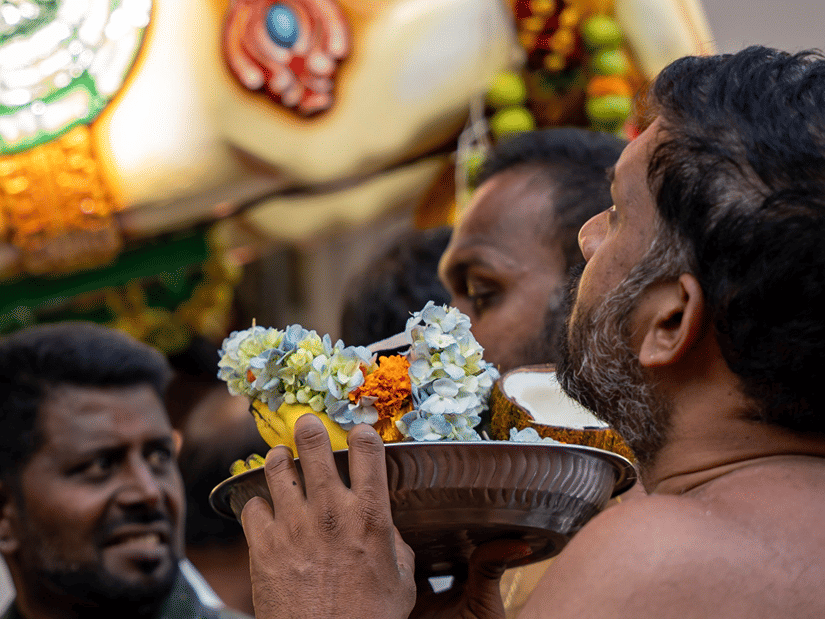 Devotees gathered around a decorated religious figure during a temple ritual, with one man leaning forward towards the offering.