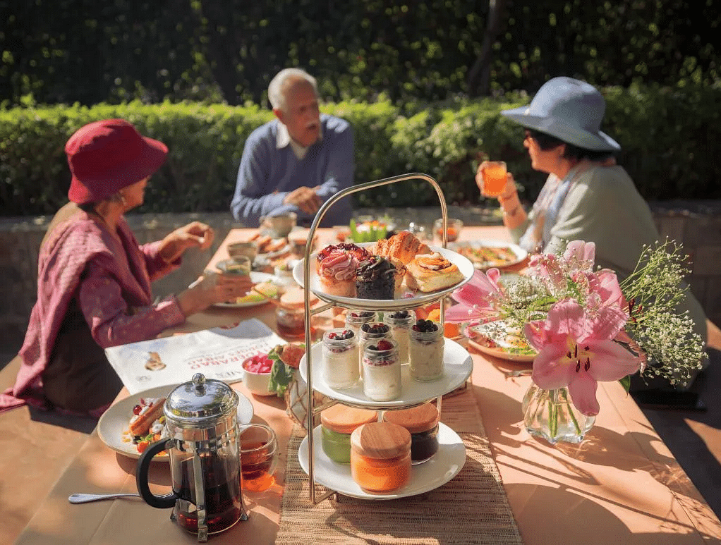A group of people in a high tea setup outdoors with a tiered tray, flowers, and teapots on the table at Karma Chalets.