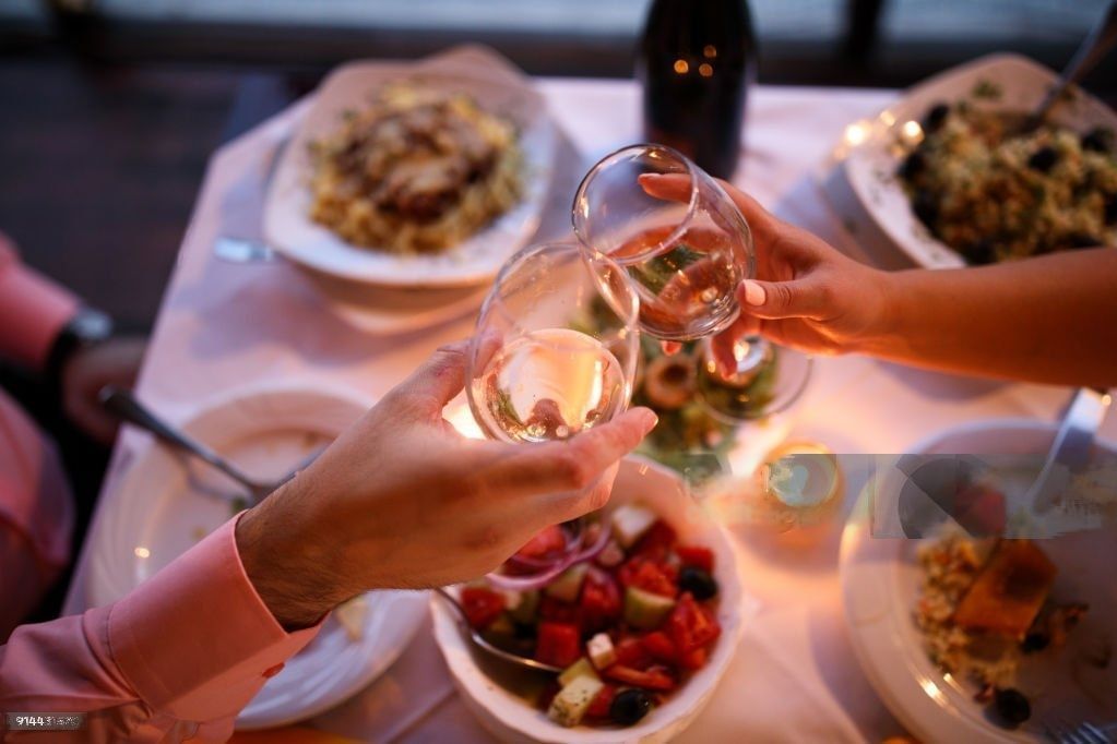 A close-up of a romantic dinner setting with two pairs of hands clinking wine glasses over a table with plates of food, candles, and a soft, warm glow.