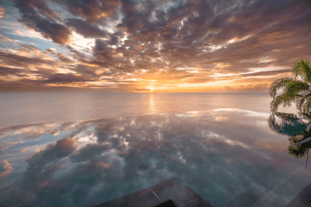 A vibrant sunset casts reflections across the still surface of an infinity pool or body of water, meeting the horizon.