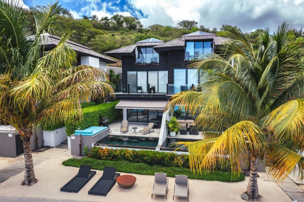Luxury beachfront villa surrounded by palm trees, featuring multiple balconies, private pool, and sunbeds.