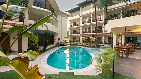 ourtyard view of a hotel in Goa featuring a central swimming pool, sun loungers, palm trees, and adjacent outdoor dining area