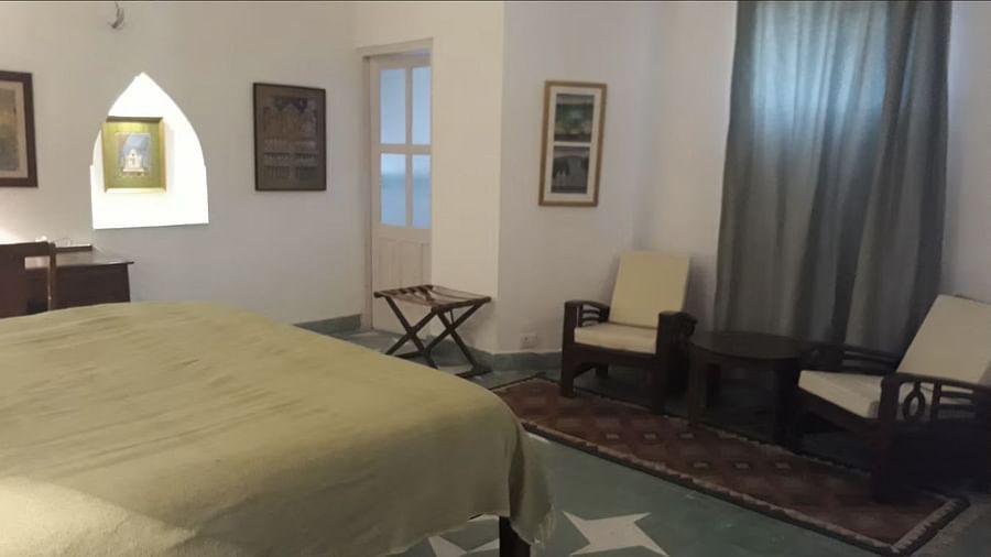 A room with a light green bed, two beige armchairs, framed artwork on the walls, and a window with sheer gray curtains, showcasing a simple and elegant design - Tijara Fort-Palace - 19th Century, Alwar.