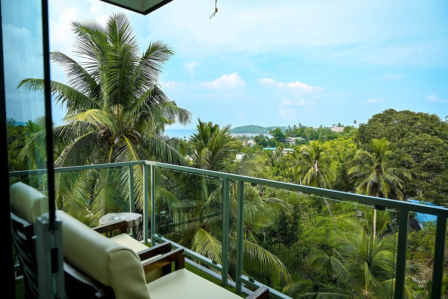 A stunning view from a hotel balcony with a glass railing, overlooking lush green palm trees and a distant view of the sea and coastline under a bright sky - Sea Hills Hotels, Sri Vijaya Puram (Port Blair)