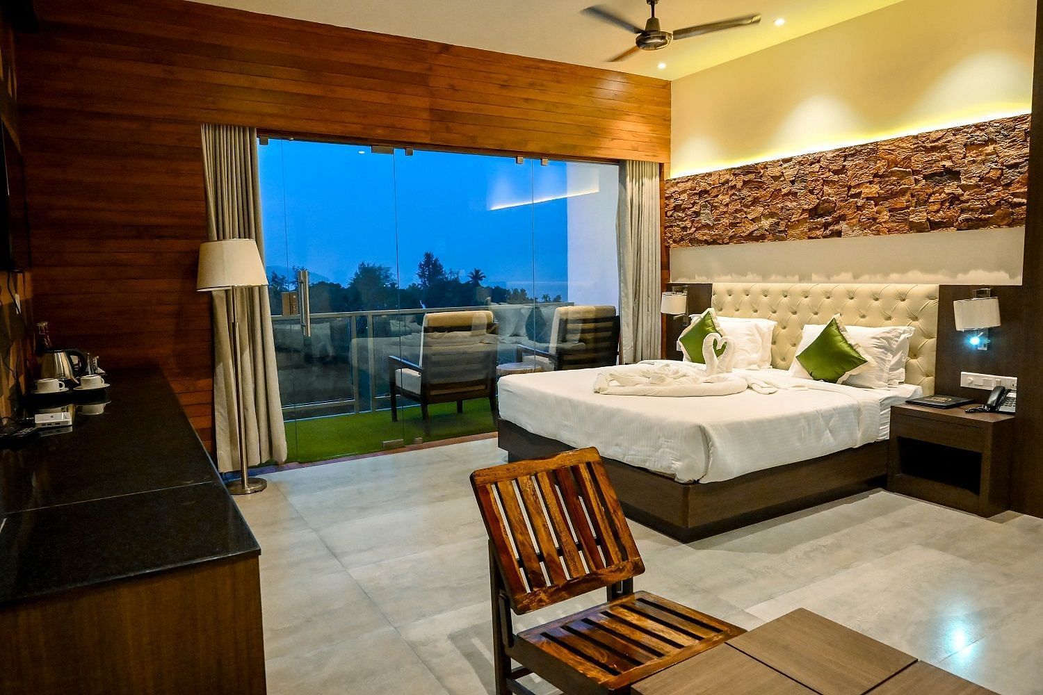 A hotel bedroom with a large bed, a comfortable armchair, and a wide view of the outdoors through a large glass door leading to a balcony - Sea Hills Hotels, Sri Vijaya Puram (Port Blair)