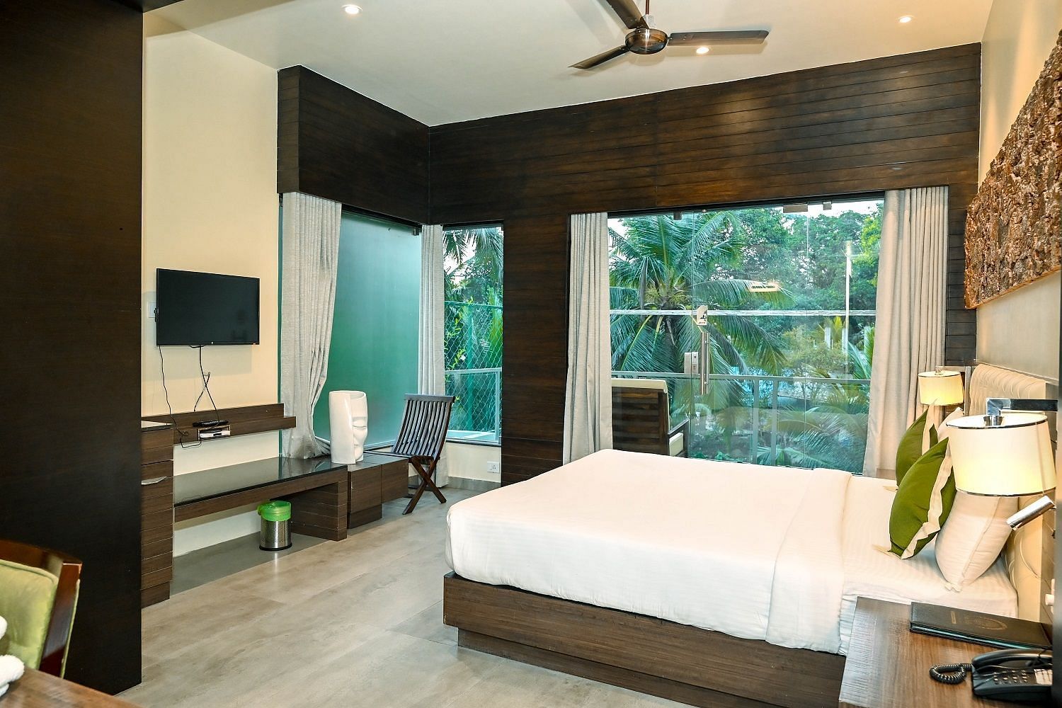 A hotel room with a large bed, a television, and a panoramic window revealing a vibrant green landscape outside, under a ceiling fan - Sea Hills Hotels, Sri Vijaya Puram (Port Blair)