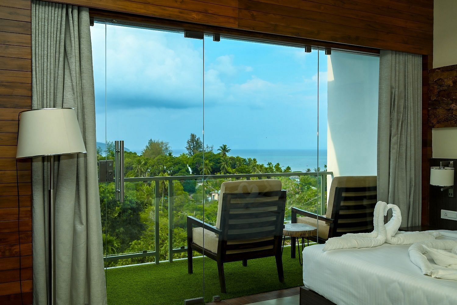 A well-appointed hotel room with a large bed, a floor lamp, and a balcony with outdoor seating offering a picturesque view of the greenery and the sea beyond - Sea Hills Hotels, Sri Vijaya Puram (Port Blair)