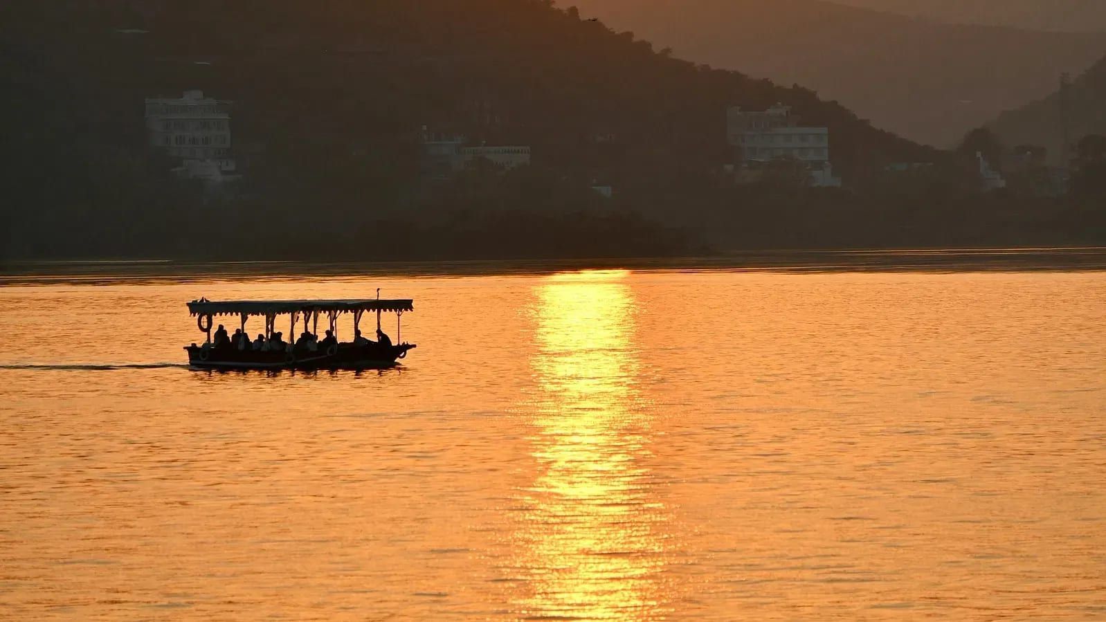 Boat silhouetted against a golden sunlight over a tranquil lake, surrounded by hills.
