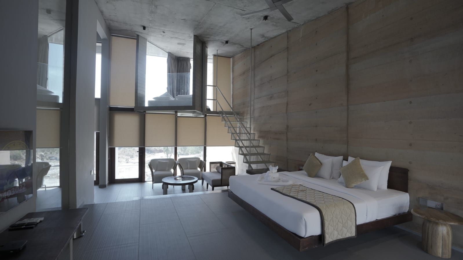 Spacious modern bedroom with tall windows, a cozy seating area, and a staircase leading to a loft-style upper section