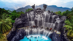 A scenic view within Black Thunder - Water Theme Park, featuring a large waterfall cascading down a rock face with a statue at the top, surrounded by lush greenery and people enjoying the water.