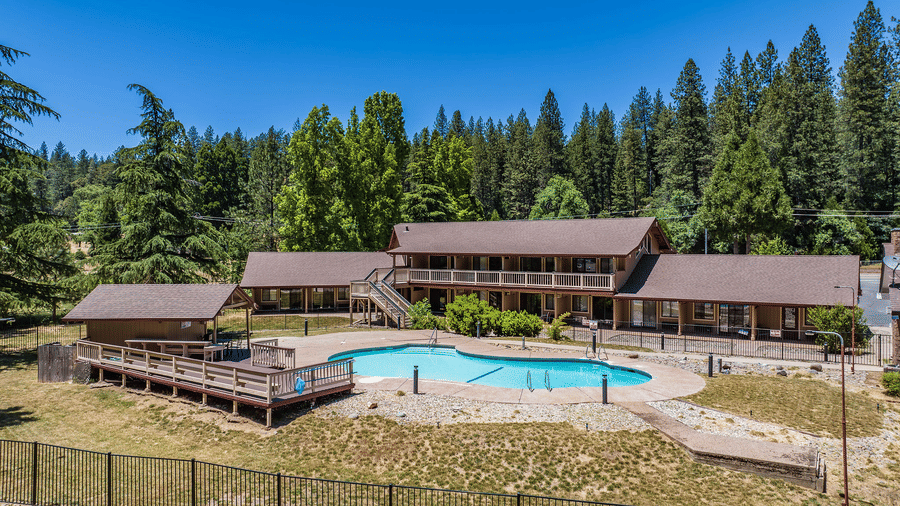 Panoramic view of Amador Hotel, one of the finest Amador County hotels, showcasing a spacious outdoor swimming pool surrounded by wooden buildings, sun deck seating, and lush pine forest.