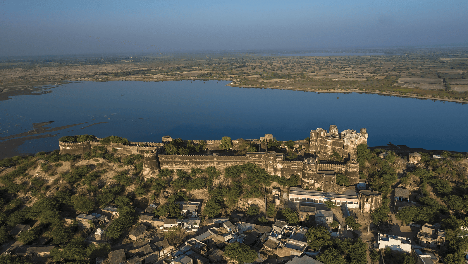 An aerial view of a sprawling fort complex on a hill, overlooking a large body of water and a hazy landscape stretching to the horizon- Shahpura Bagh.