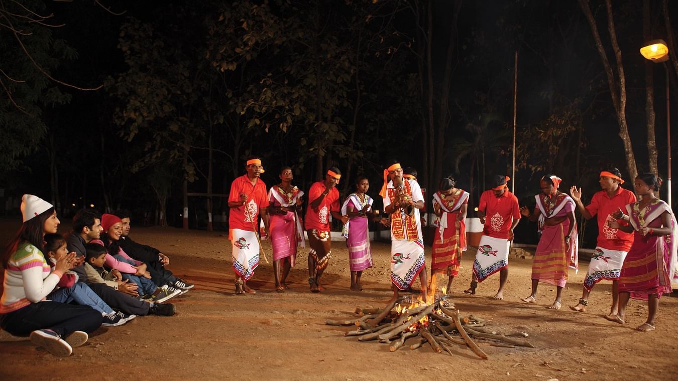 A group of people sitting near to a camp fire, seen clapping for a group of performers in traditional clothes at night