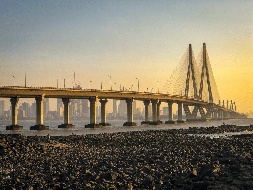 Golden-hour view of the Bandra–Worli Sea Link in Mumbai, a long cable-stayed bridge stretching across the Arabian Sea, with city skyscrapers in the background and a rocky shoreline in the foreground.