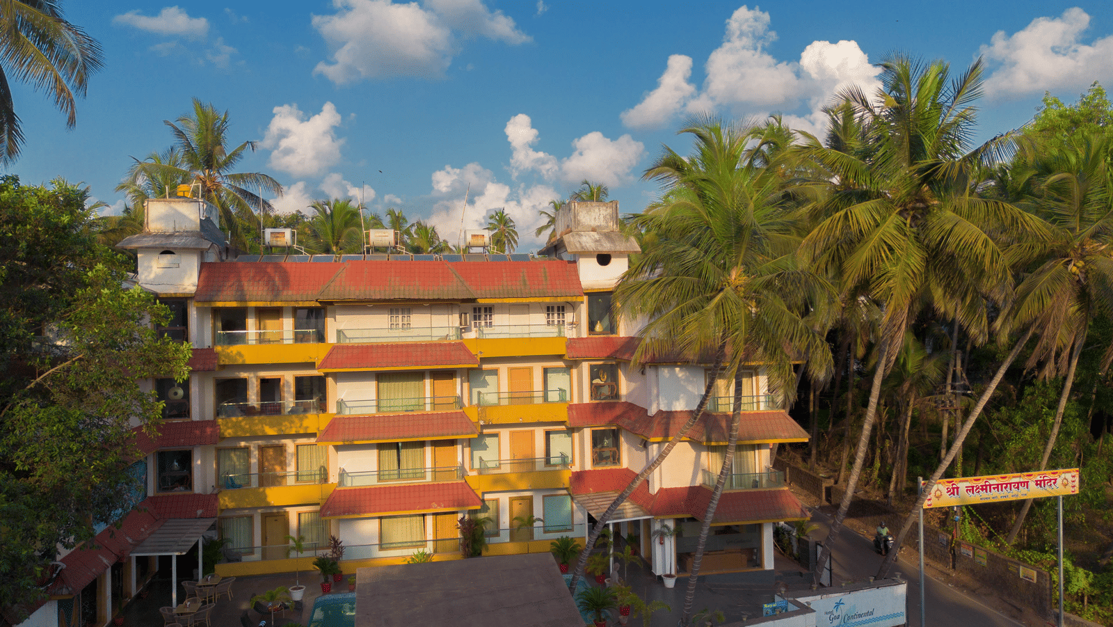 A colourful, low-rise hotel complex with many windows, surrounded by lush green palm trees under a bright sky - Perfectstayz Premium Goa Continental