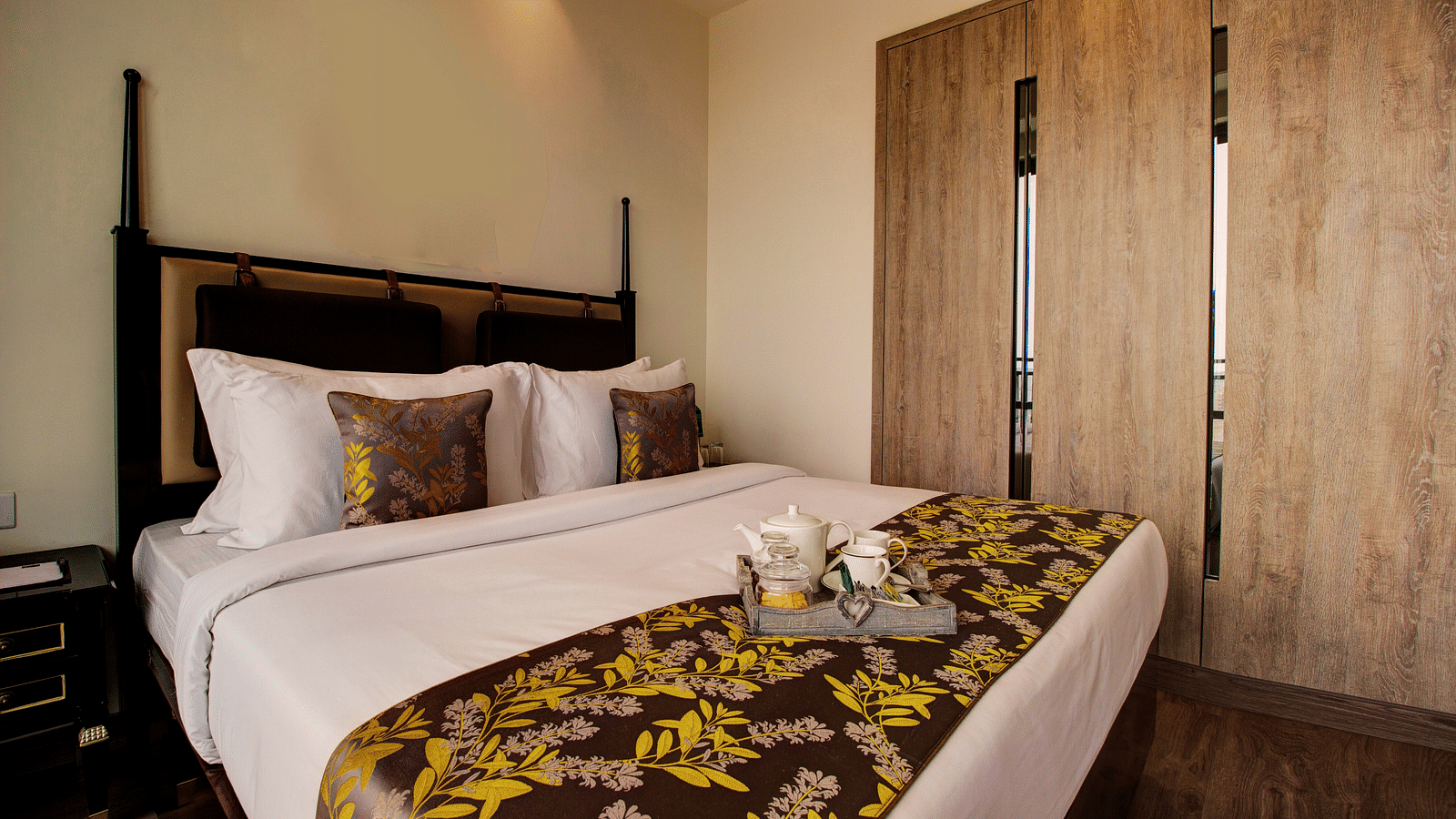 A plush bed on with pillows and cushions and a tray containing cutlery for hot beverages sitting on the bed runner with a large closet in the background
