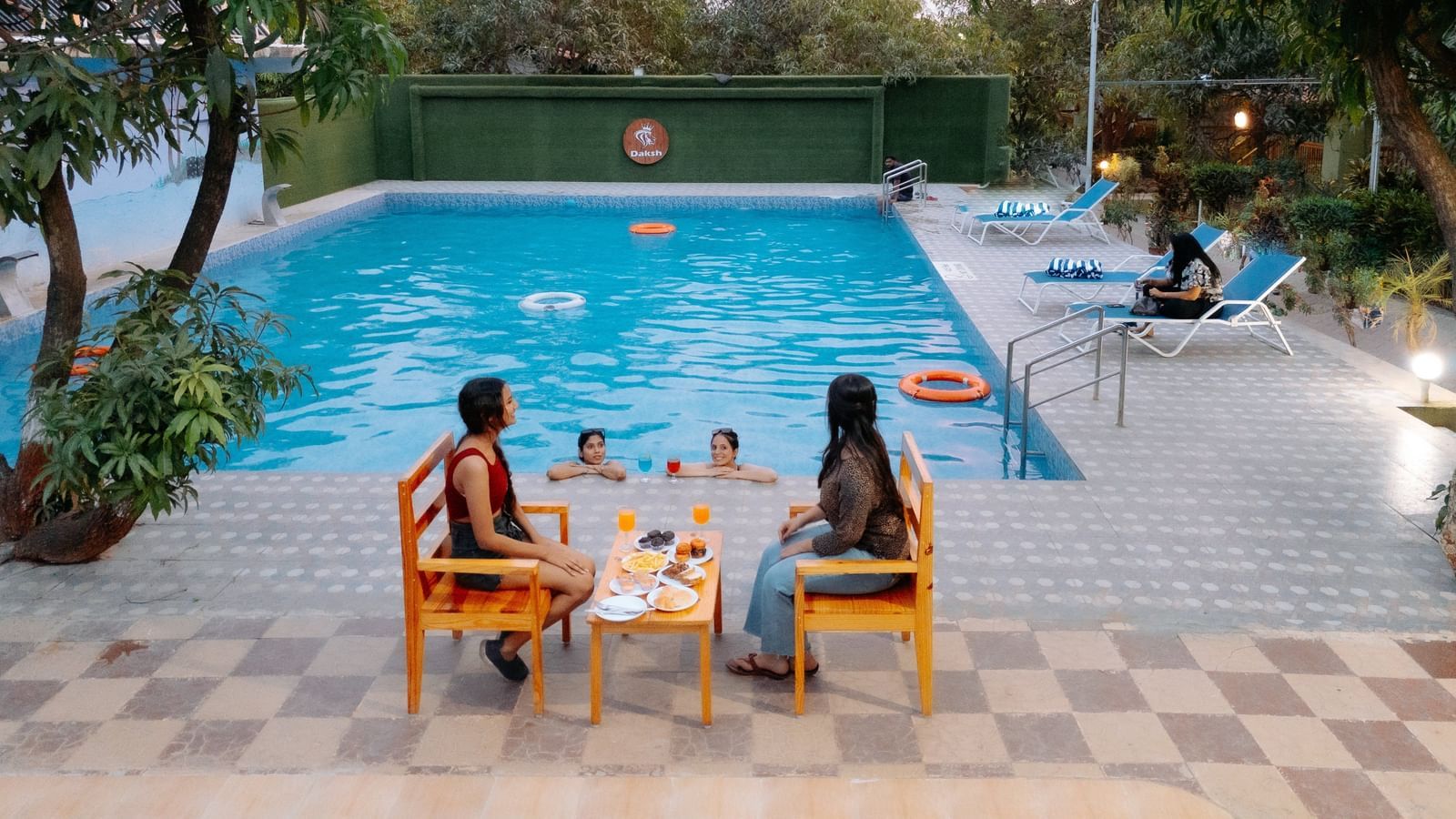 Two people seated at a table near a swimming pool, while others swim and relax around the water at Daksh Resort & Amusement Park.