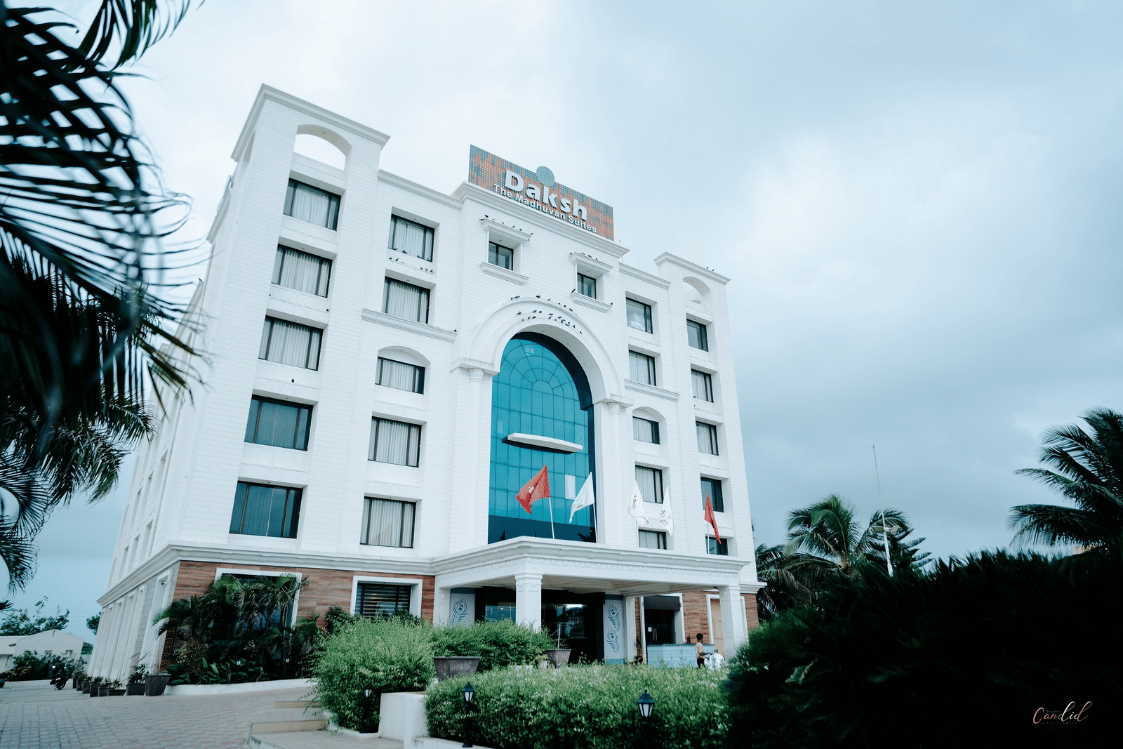 Large, modern white hotel building exterior on a slightly overcast day. The main entrance features a large arched blue glass window at Daksh The Madhuvan Suites, Dwarka.