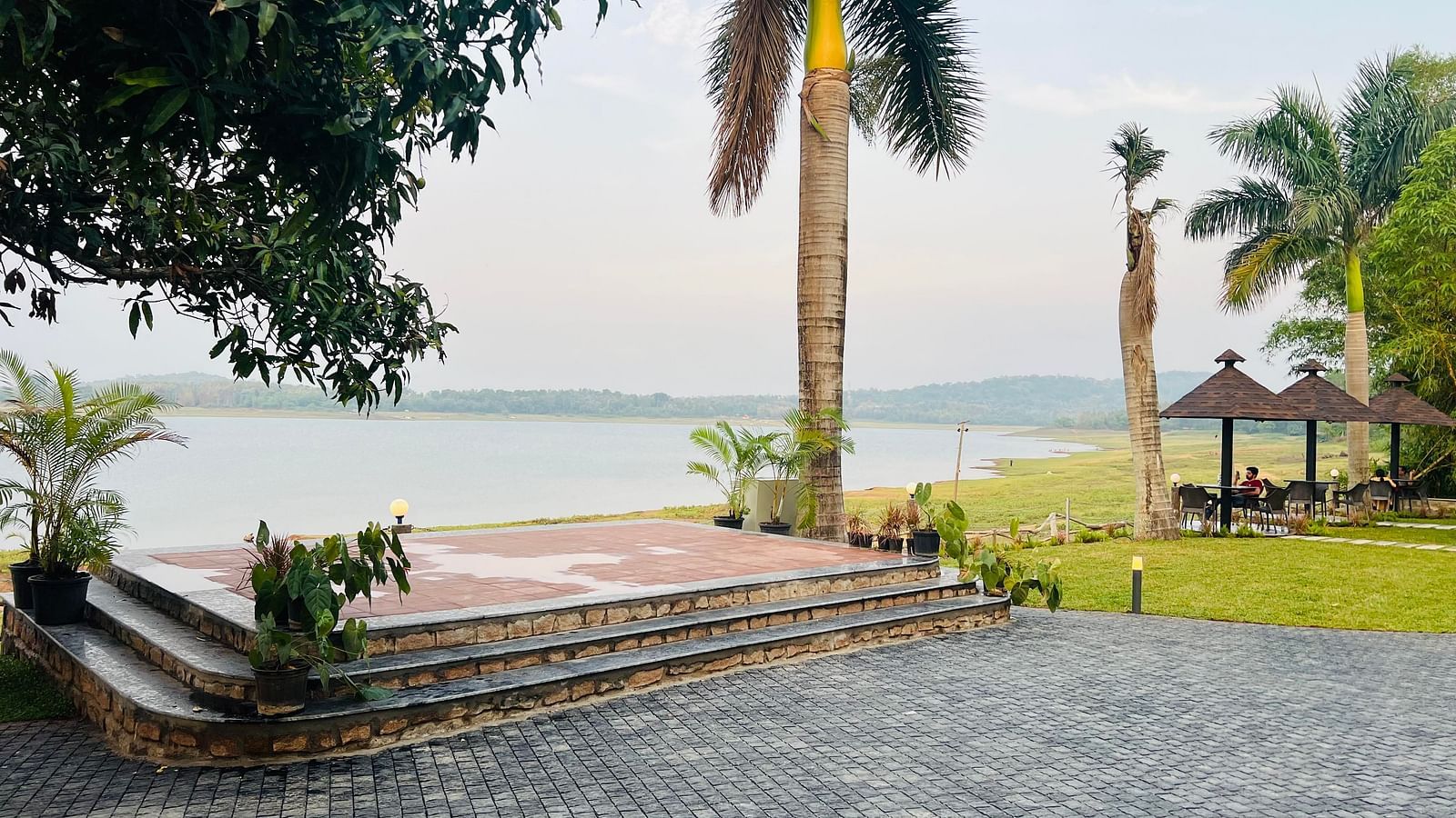 View of a lakeside area with stone steps, palm trees, and a paved pathway. A gazebo with a thatched roof sits near the water, with grassy areas and distant hills visible across the lake
