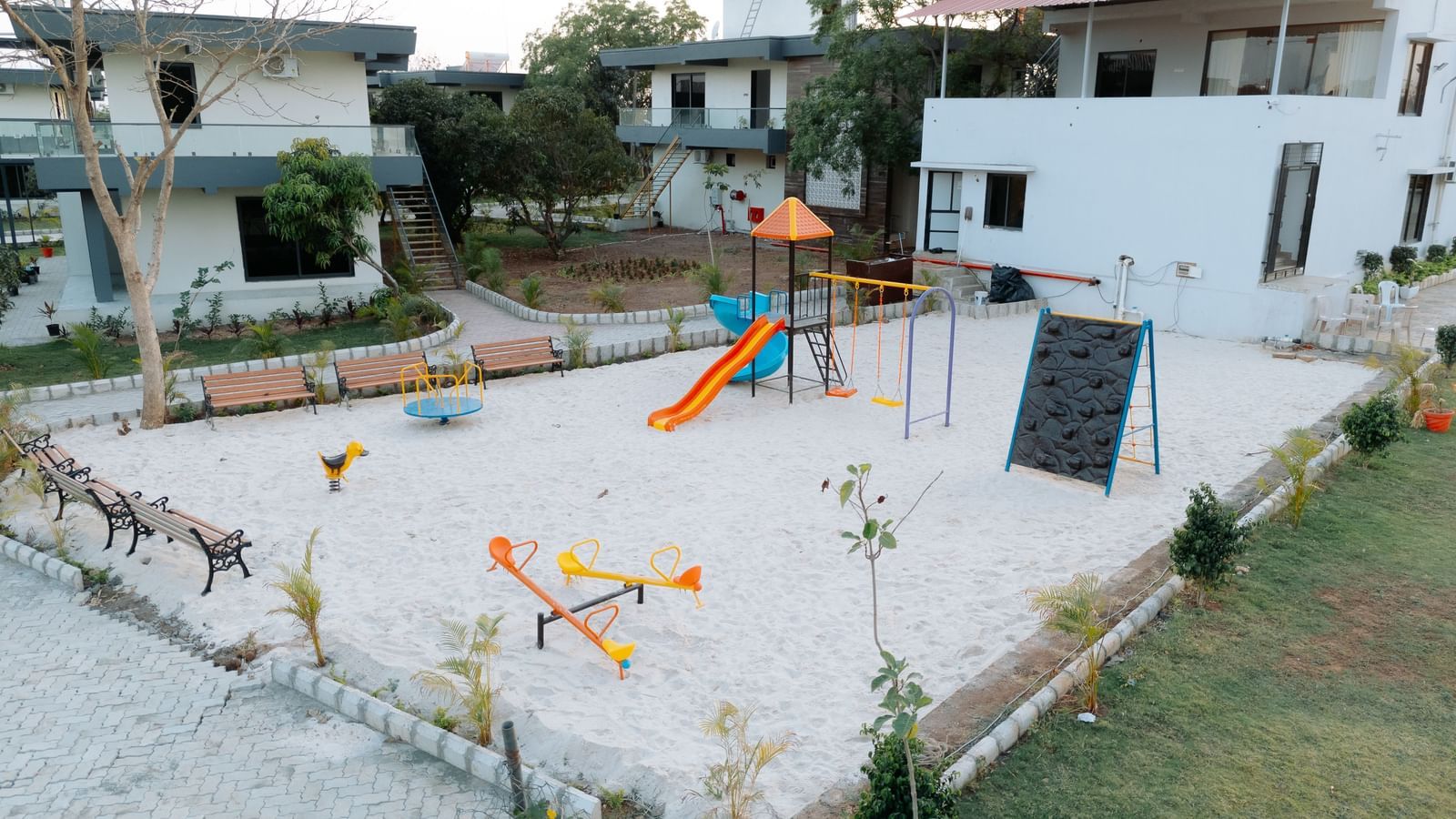 The kids' play area of Daksh Eden Greenz in Sasan Gir, features slides, swings, seesaws and more placed in a sand bottomed area with buildings and trees in the background.