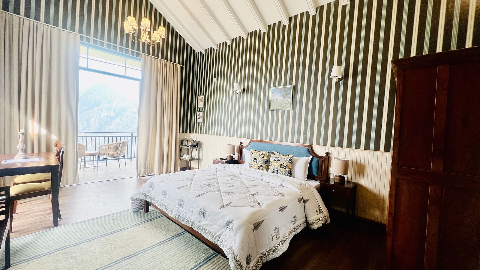 Spacious hotel room featuring a wooden bed with white and gray bedding, striped walls, and a panoramic window with mountain views | Faraway Cottages, Auli