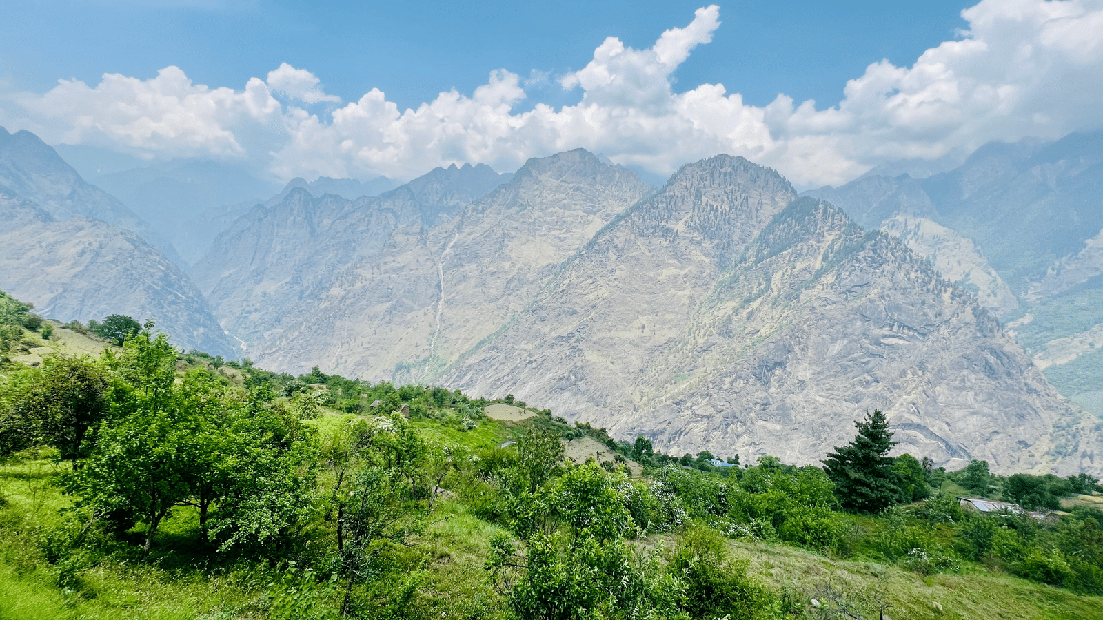 Lush green hills and trees with towering mountain peaks under a bright, partly cloudy sky.