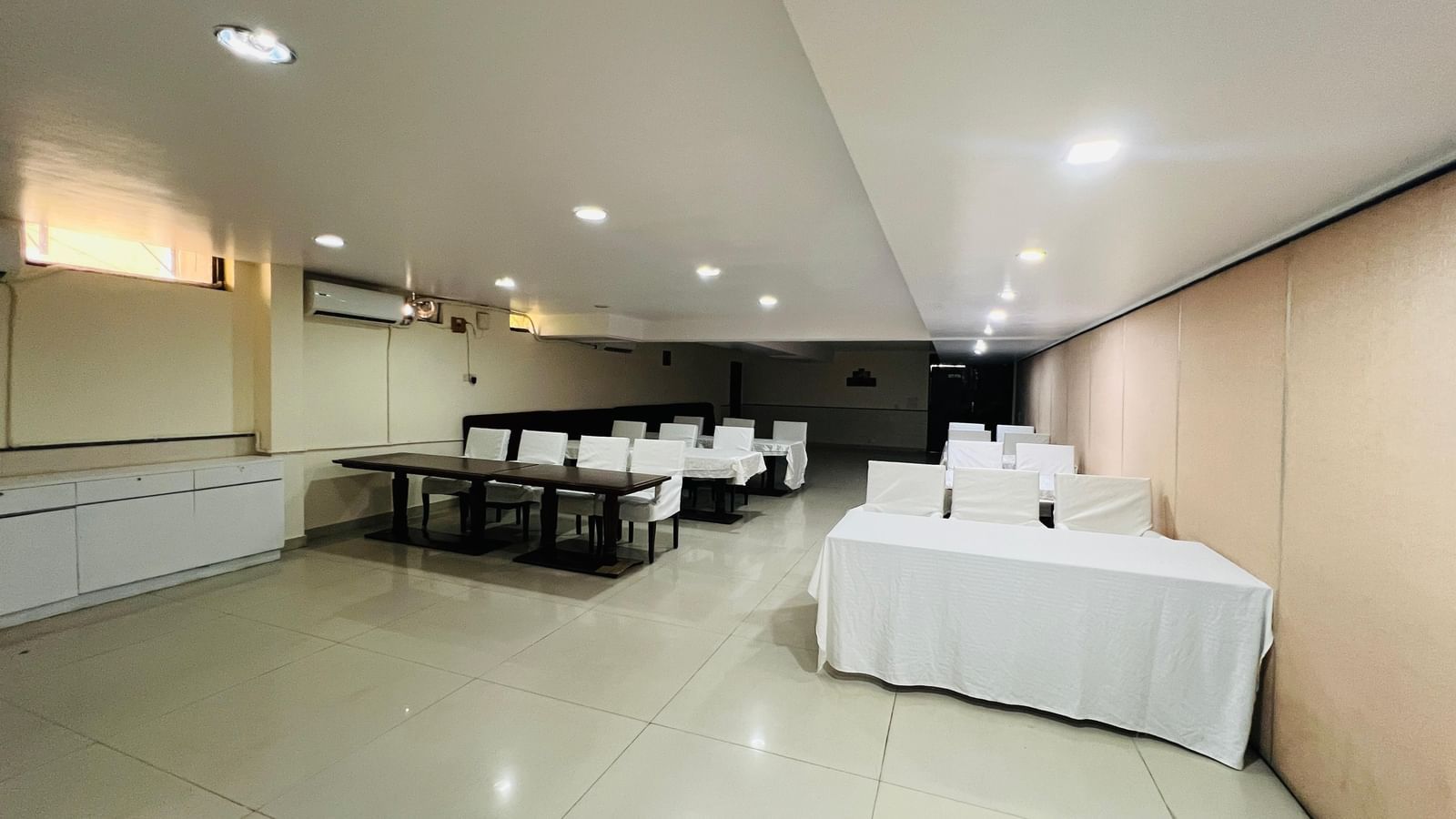  A hotel conference room with long tables and white chairs arranged for an event, featuring modern lighting and light-colored walls. | Hotel Aadhar