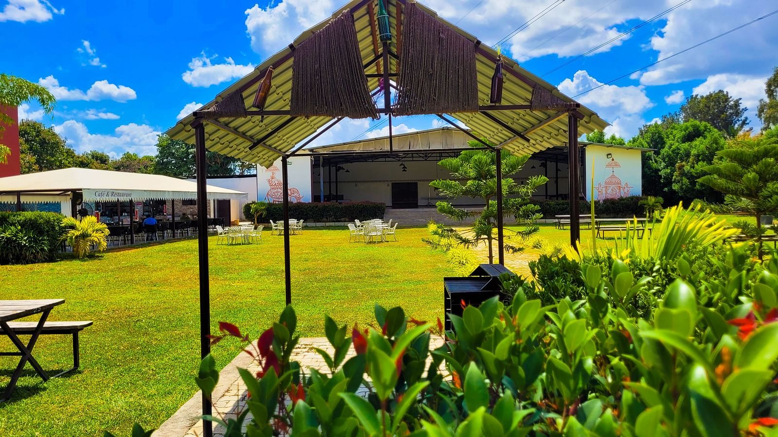 Covered area with tables lined under a beige canopy amidst lush greenery. | Vistar Resort, Bengaluru