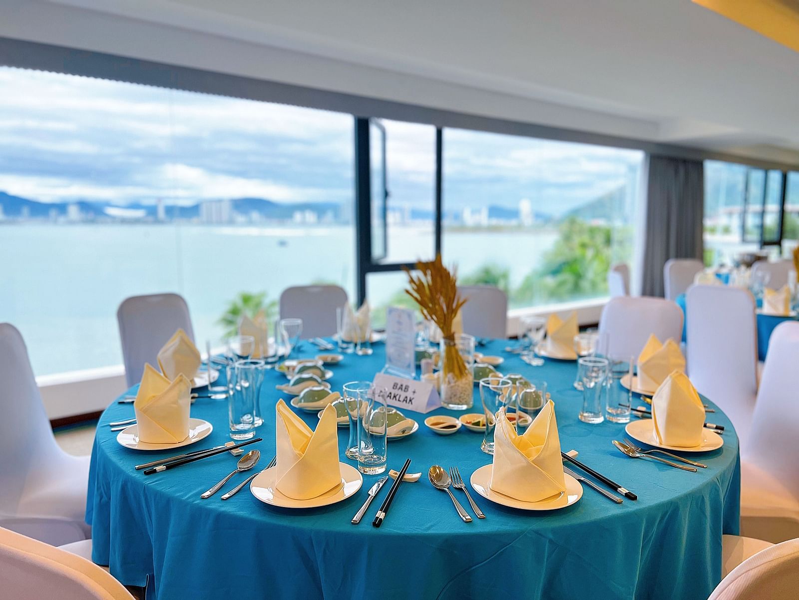 A close up shot of a round table covered in a blue cloth with neatly founded napkins on plates - Alibu Resort