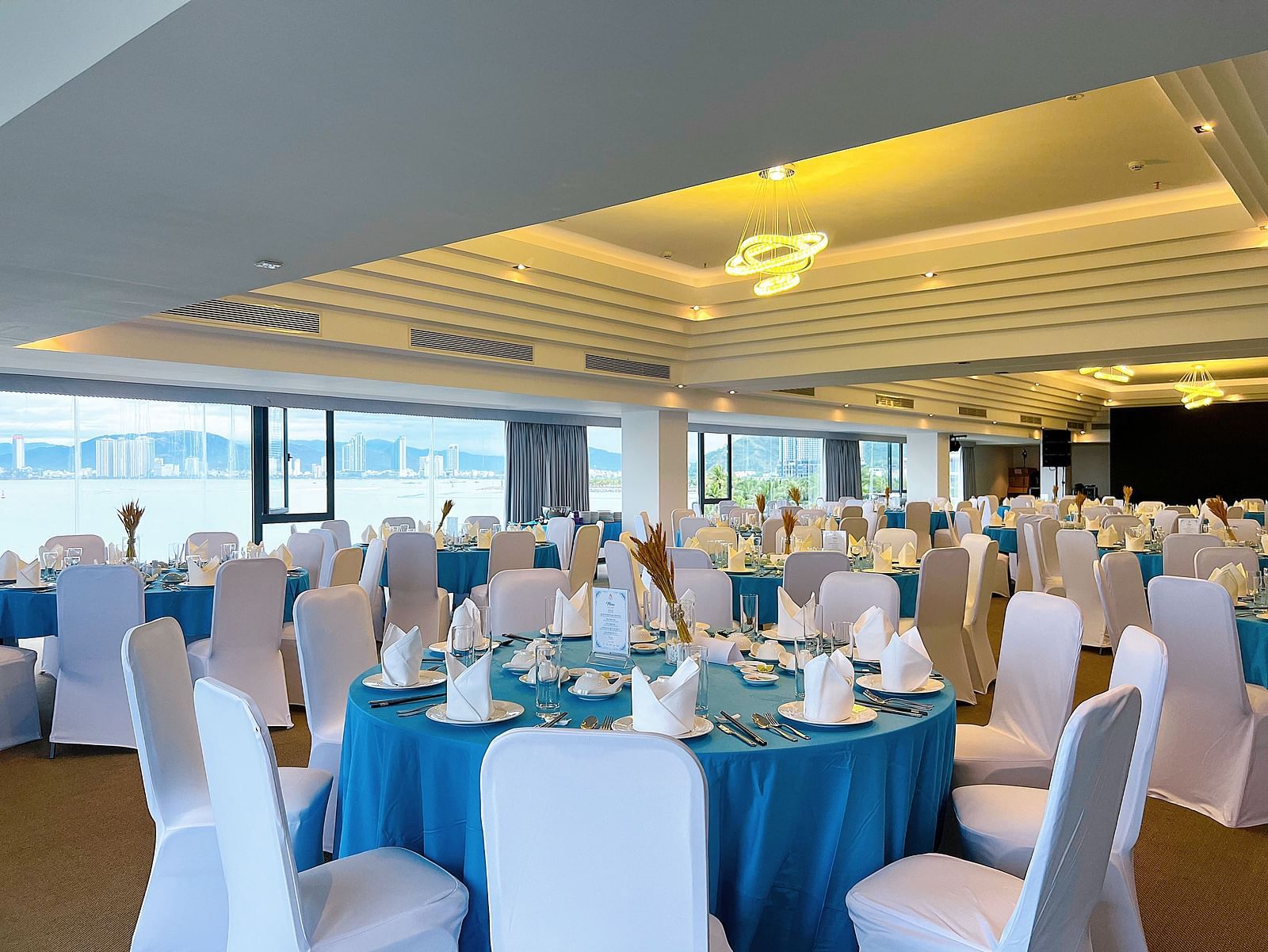 A meeting hall with blue round tables, ambient ceiling lighting and a view of the Nha Trang Bay - Alibu Resort