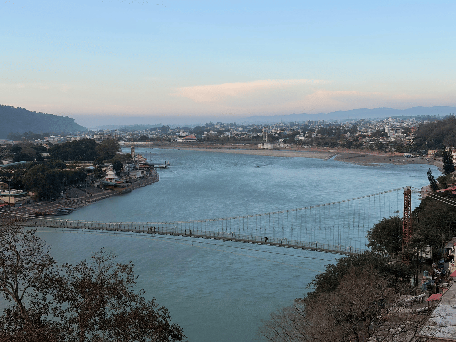 A high-angle view of a long suspension bridge stretching across a wide river at dusk.