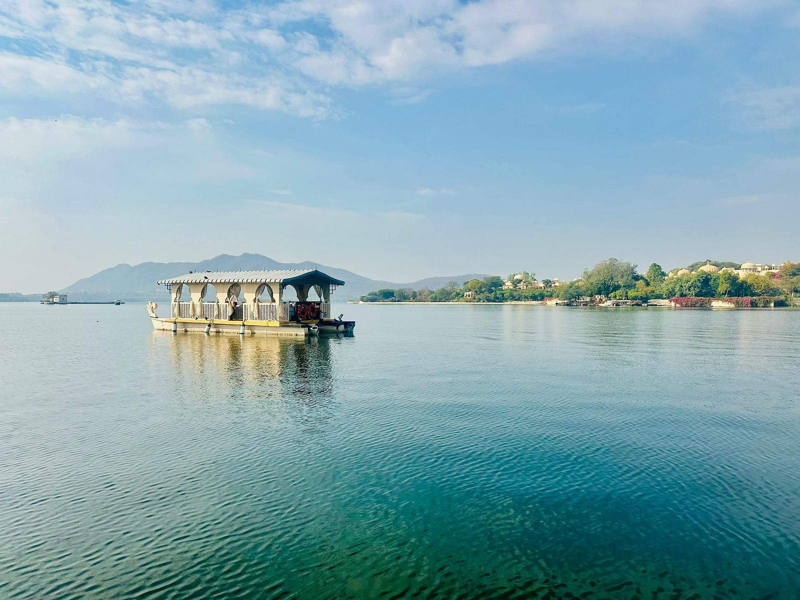 A traditional houseboat floats peacefully on the still, blue waters of a lake, with distant green hills under a clear sky.
