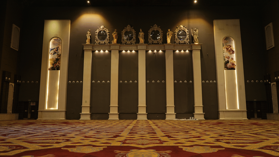 Formal stage backdrop with vertical panels, subtle lighting, and framed portraits or seals displayed high up on the wall at Hotel Hukam's Lalit Mahal.