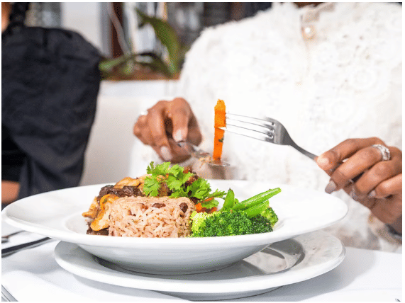 A person holding a fork and knife while eating a plate of rice with broccoli and vegetables at the restaurant of S Hotel Kingston.