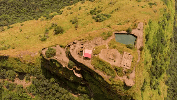 Aerial view of a hilltop fort with ruins and natural reservoirs surrounded by lush landscape.