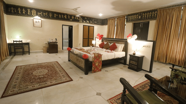 A spacious bedroom with a large bed decorated with towel art, bedside tables with lamps, a seating area with chairs and a table, a wall-mounted air conditioner, and large curtained windows at Infinity Resorts Bandhavgarh.