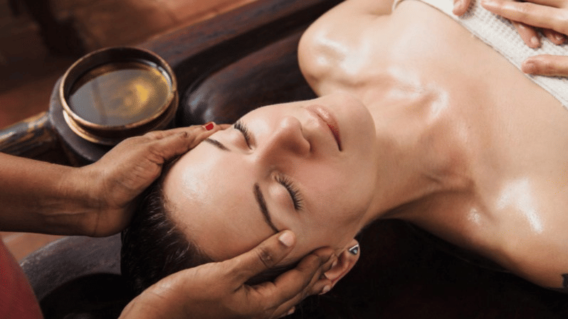 A woman lying on her back with her eyes closed receiving an Ayurvedic head and face massage with oil