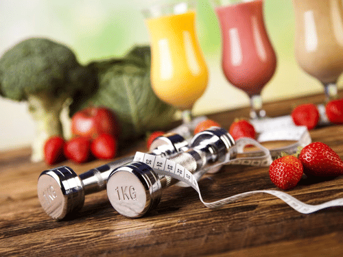 Dumbbells and measuring tape on a table with a variety of fresh smoothies and vegetables in the background, promoting a healthy lifestyle.