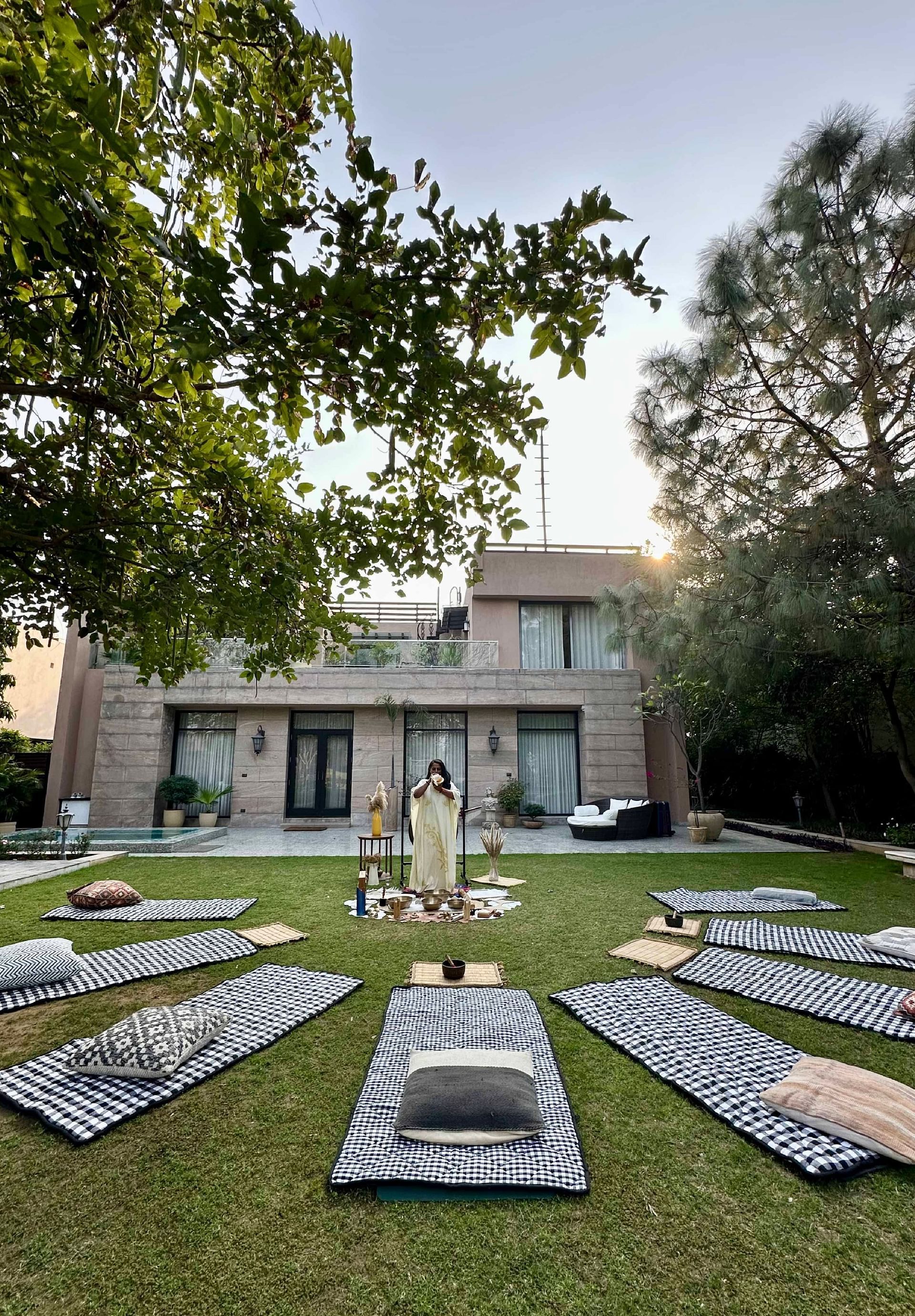 A person blowing a conch and standing between a lawn area that is laid with yoga mats, turkey towels and partially canopied by a tree - Karma Chalets
