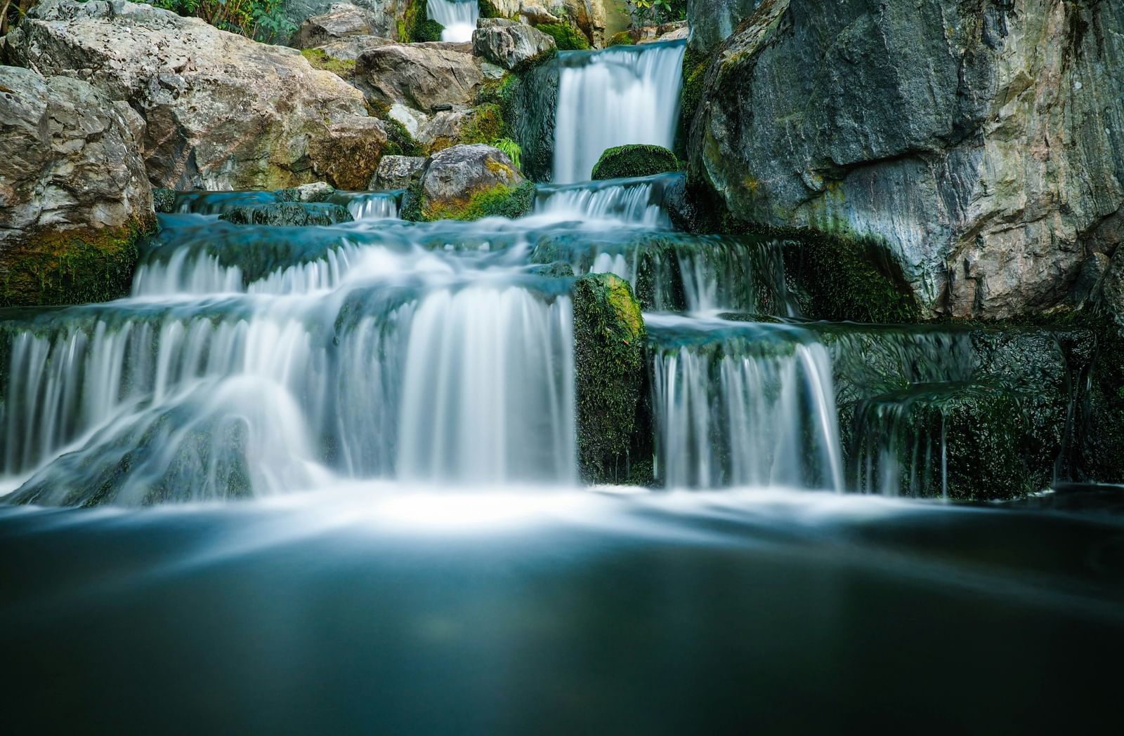 An eye-level view of a series of small waterfalls over smooth, moss-covered rocks, with a long-exposure effect blurring the water.
