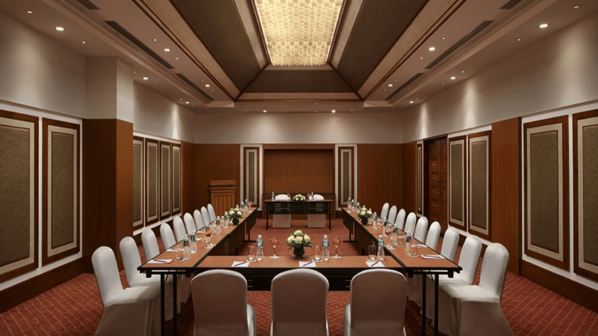 An overview of a U-shaped seating arrangement with white chairs, spot lights above on the sides and chandelier in the centre of the roof inside a banquet gall in Goa at Caravela Beach Resort.