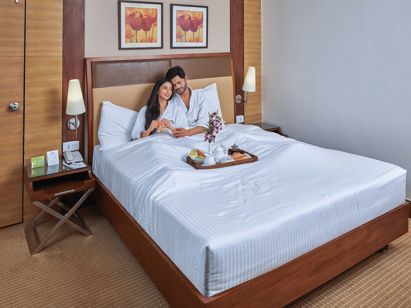 A couple sitting on a bed in a hotel room decorated for a honeymoon package, featuring food on the bed