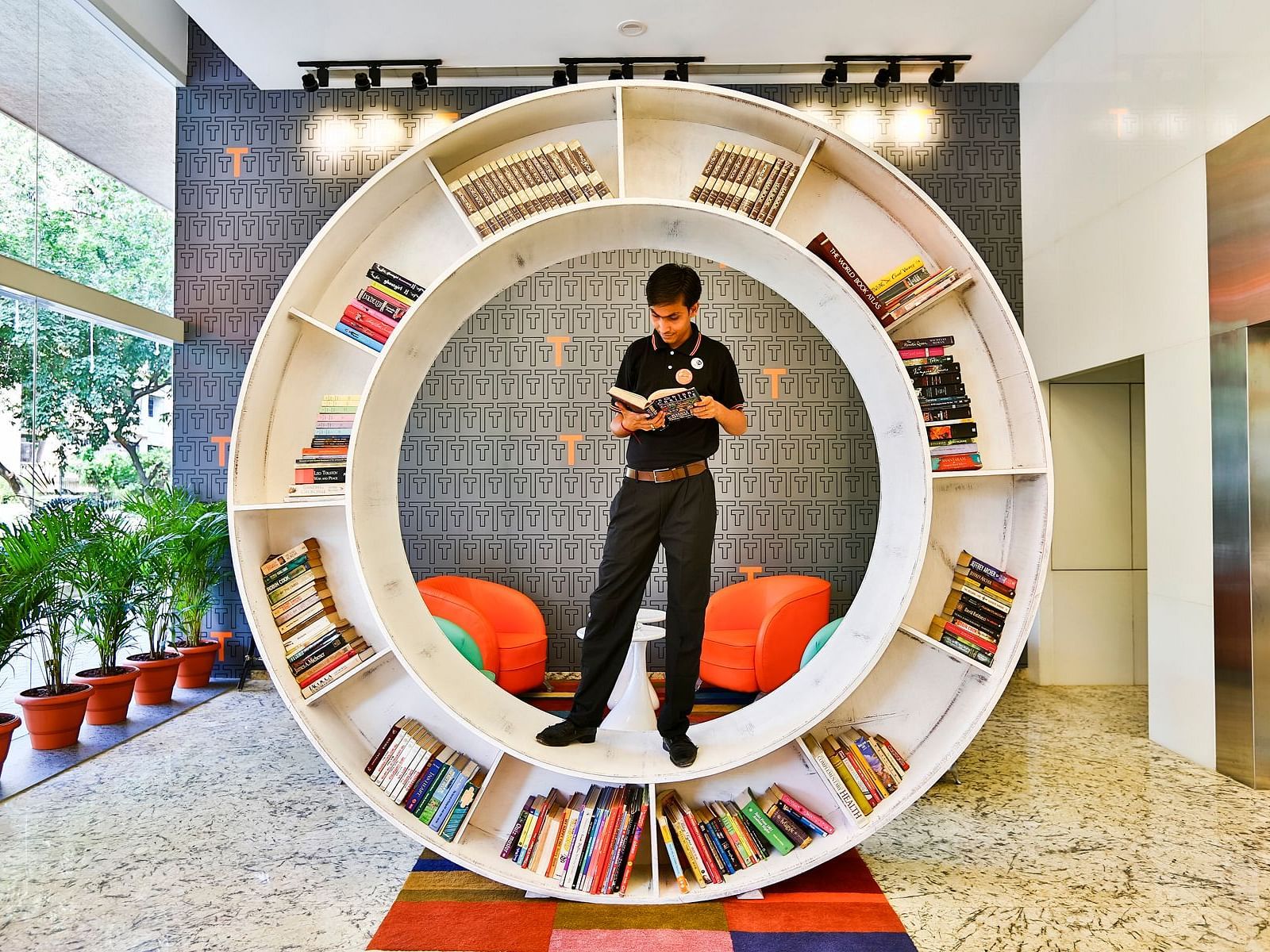 A hotel employee stands inside a large circular bookshelf, reading a book. The bookshelf is filled with books and is located in the lobby of T24 Residency.
