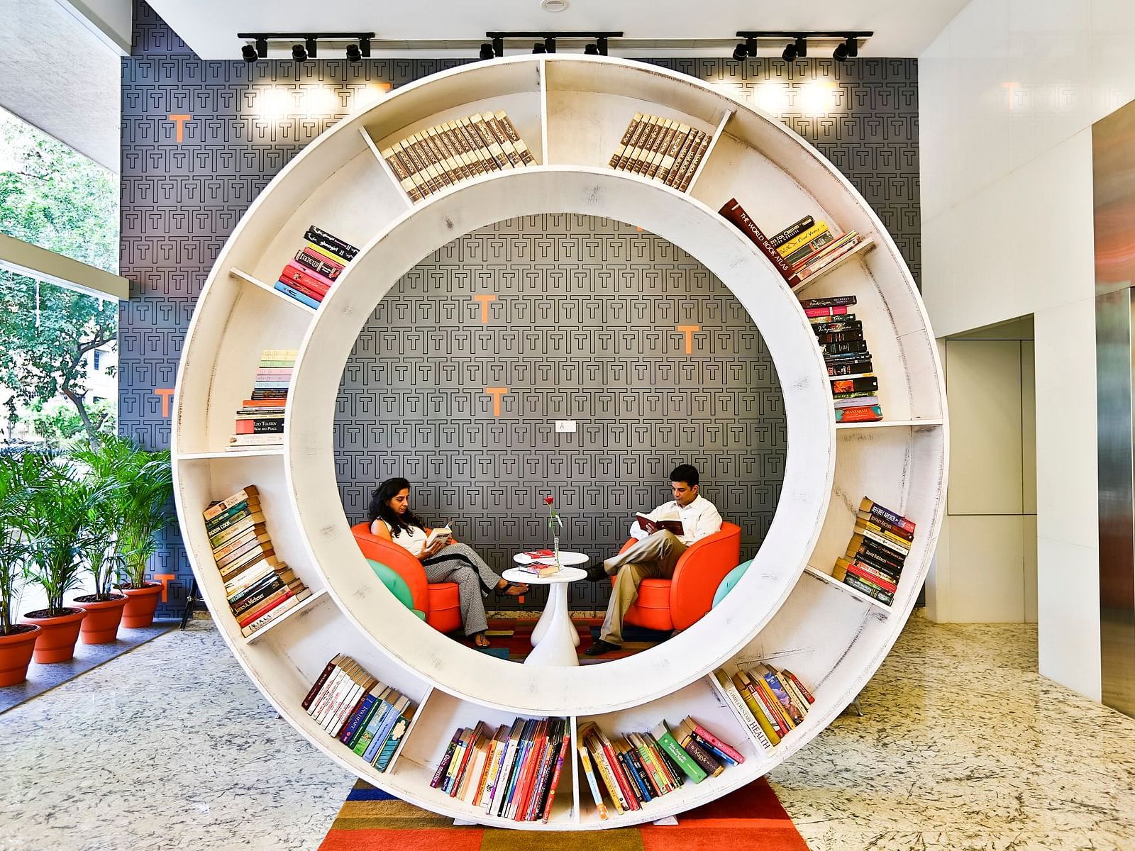 Two people sit in comfortable orange chairs inside a circular bookshelf in the lobby of the T24 Residency hotel in Mumbai, India.