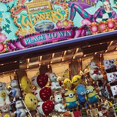 A prize display at a fair or arcade shows many stuffed animals and other small toys hanging from hooks beneath a brightly decorated sign.