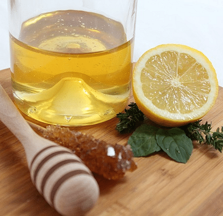 An image featuring a cup of honey, half a lime, herbs, a big honey dipper decoratively placed on a wooden plate.