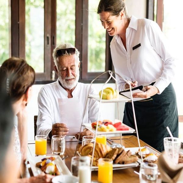 A smiling staff standing next to a man in white shirt viewing a sheet, sitting at the head of a table set with pastries and other delicacies.